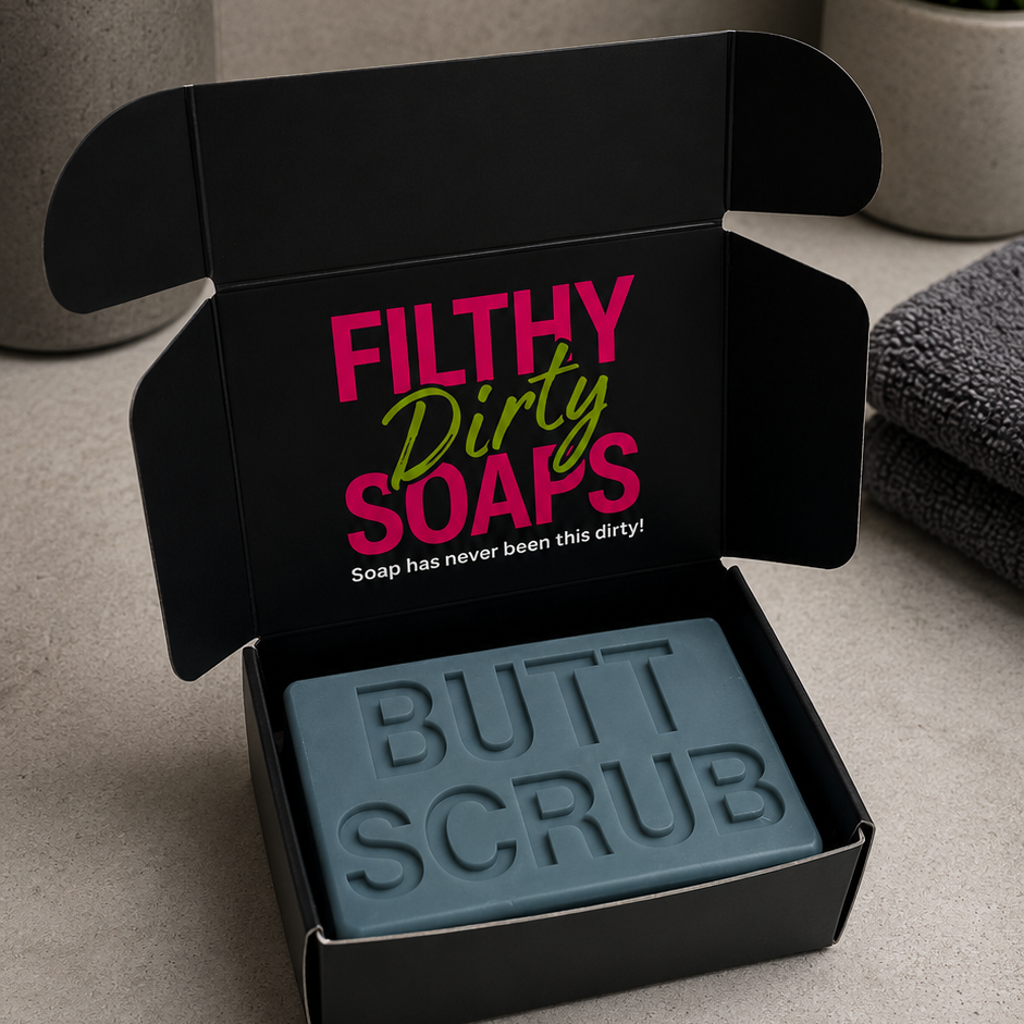 A dark gray bar of 'Butt Scrub' soap in an open black gift box with the brand name 'Filthy Dirty Soaps' printed on the inside lid. 