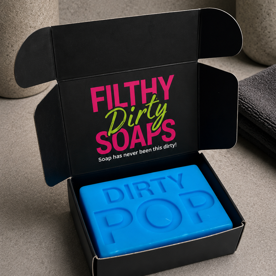 A bright blue bar of soap labeled 'DIRTY POP' sits inside an open black gift box with the words 'FILTHY Dirty SOAPS' printed on the inside lid. 