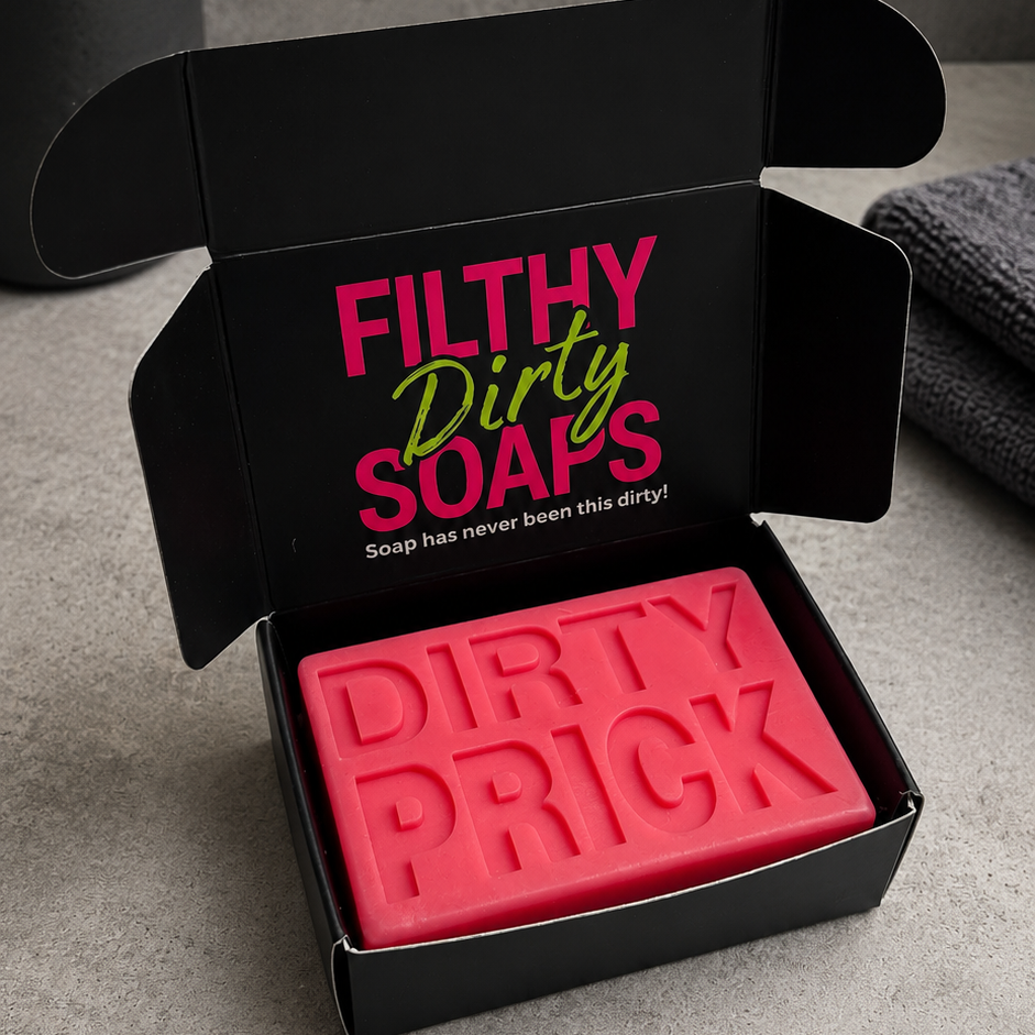 A red bar soap bar embossed with the words 'DIRTY PRICK' sits inside an open black gift box. The box lid features the brand name 'Filthy Dirty Soaps' in pink and green lettering.