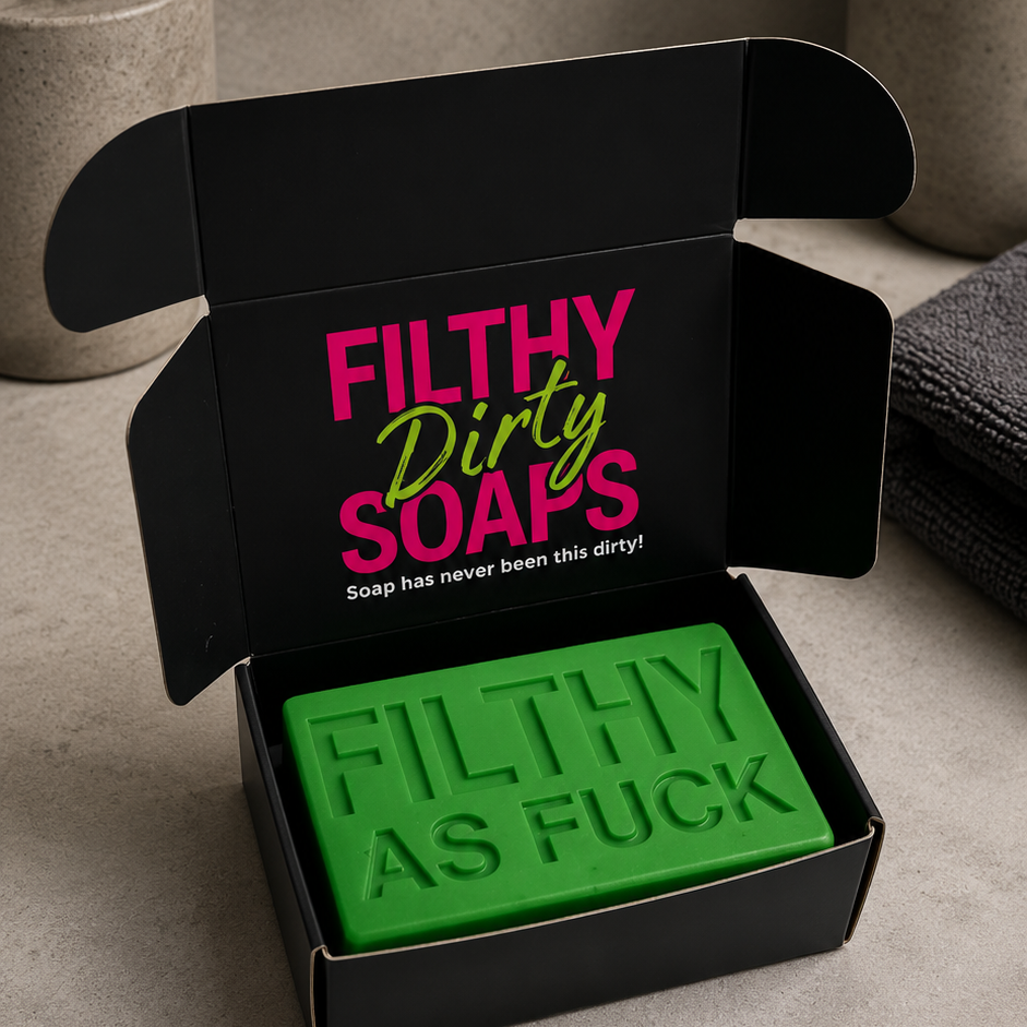 A green rectangular bar of soap embossed with 'FILTHY AS FUCK' sits inside an open black gift box. The inside lid has pink and green text reading 'FILTHY Dirty SOAPS' with a smaller tagline.