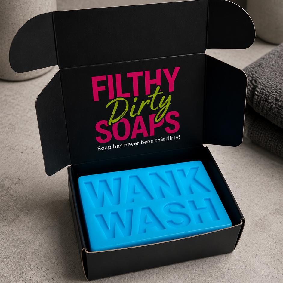 A bright blue bar of soap embossed with 'WANK WASH' sits inside an open black gift box. The box lid features the text 'FILTHY Dirty SOAPS' in pink and green.