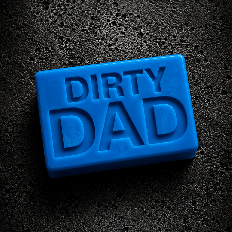 A bright blue rectangular soap bar embossed with the words 'DIRTY DAD' in raised lettering, photographed on a dark textured surface.