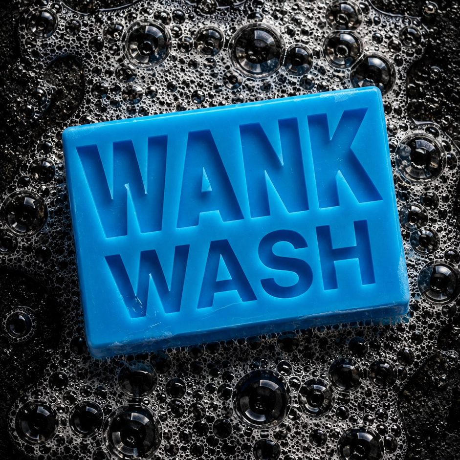 A bright blue rectangular bar of soap embossed with the words 'WANK WASH' in large letters, resting on a bed of dark soapy foam against a black background.